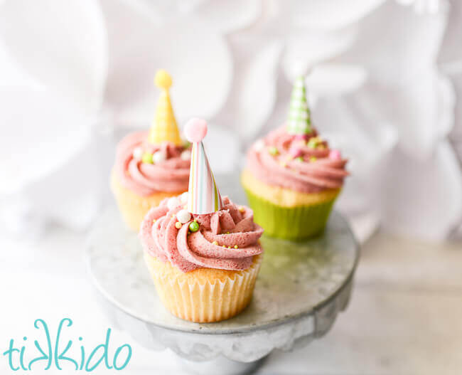 three vanila cupcakes with pink raspberry buttercream icing and topped with miniature party hat cupcake toppers, sitting on a galvanized metal cake stand.
