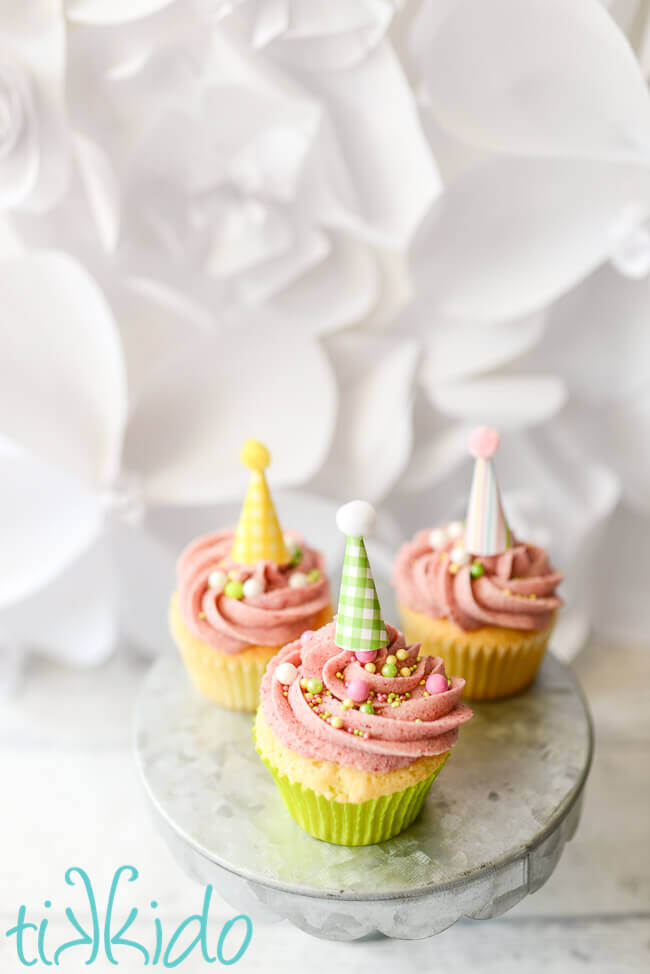 three vanilla cupcakes topped with pink raspberry buttercream, sprinkles, and miniature party hat cupcake toppers, on a galvanized metal cake stand and white backdrop.