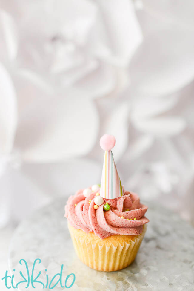 Single vanilla cupcake with pink raspberry buttercream icing, sprinkles, and a striped miniature party hat cupcake topper.