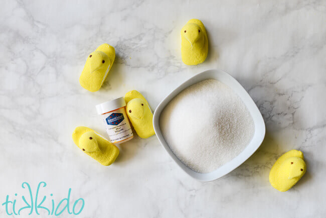 Five yellow peeps and a container of powdered food coloring next to a white bowl of sugar, on a white marble background.