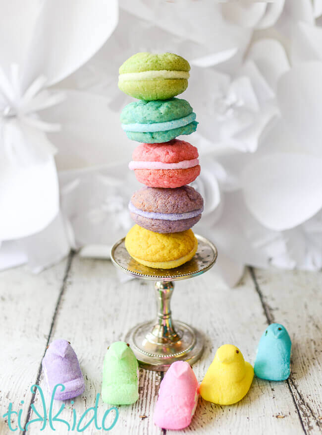 Five Peeps Whoopie pie sandwich cookies staked on top of each other on a tiny metal cake stand, surrounded by five Peeps candies in five different colors.