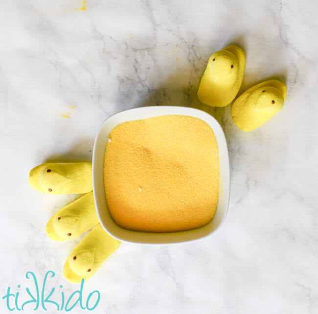 Five yellow peeps arranged around a white bowl full of yellow sugar, on a white marble background.