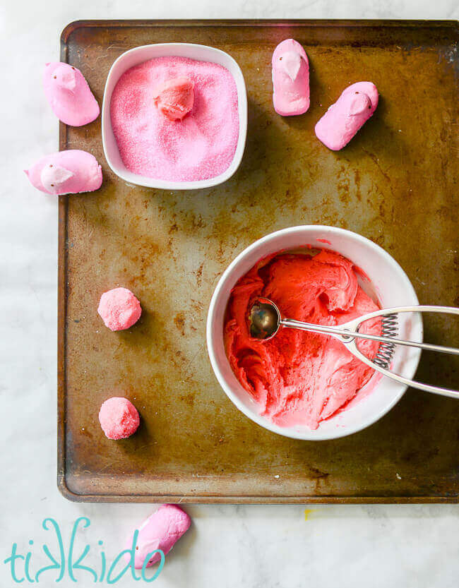 Baking sheet on white marble background, with bowl of pink sugar cookie dough, bowl of pink sugar crystals, cookie dough rolled into balls and rolled in the pink sugar, and four pink peeps.