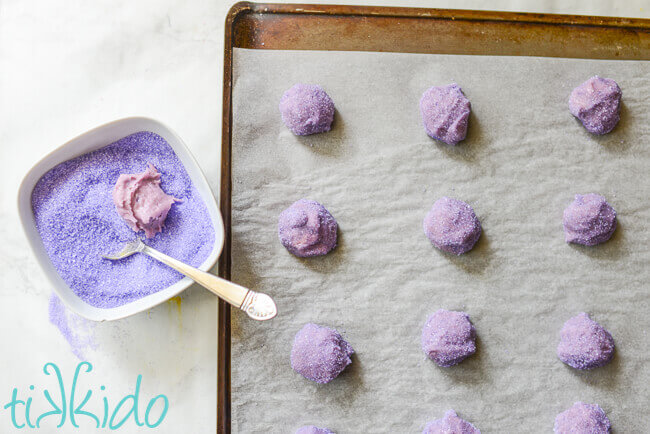 Baking sheet with parchment paper covered in purple sugar cookie dough balls rolled in purple sugar, bowl of purple sugar and purple dough ball and spoon beside the baking sheet.