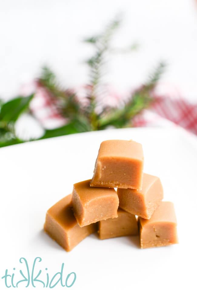 Pieces of homemade penuche fudge stacked on a white plate, with evergreen and holly branches in the background.