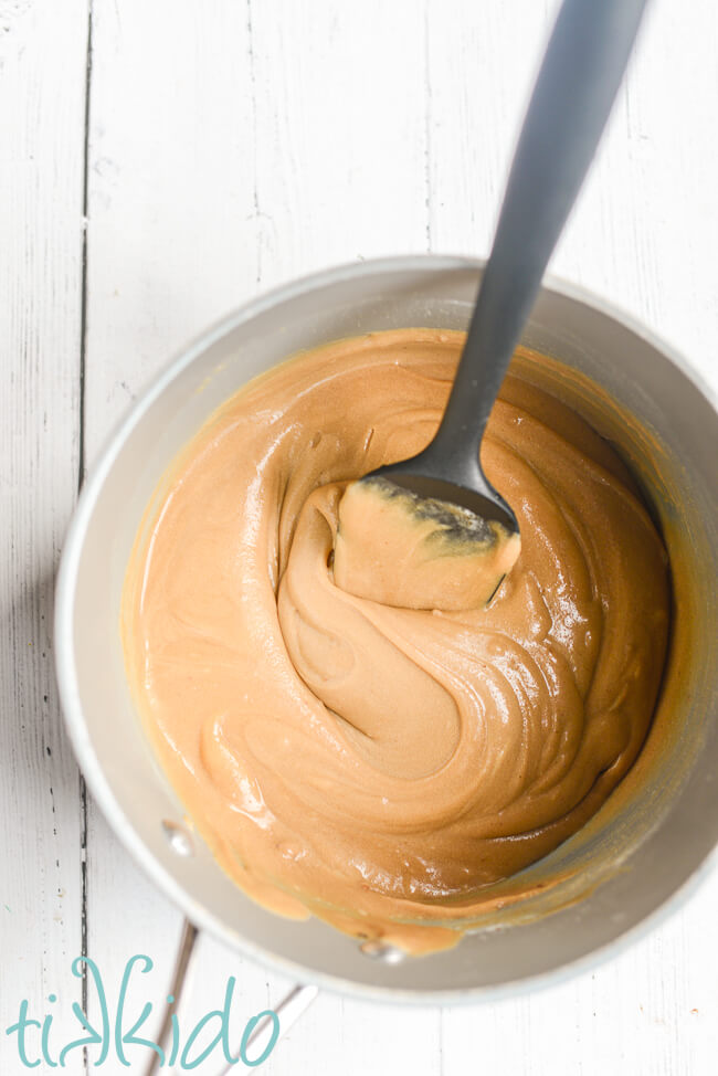 Penuche fudge ingredients stirred together with a black spatula in a large saucepan sitting on a white wooden surface.