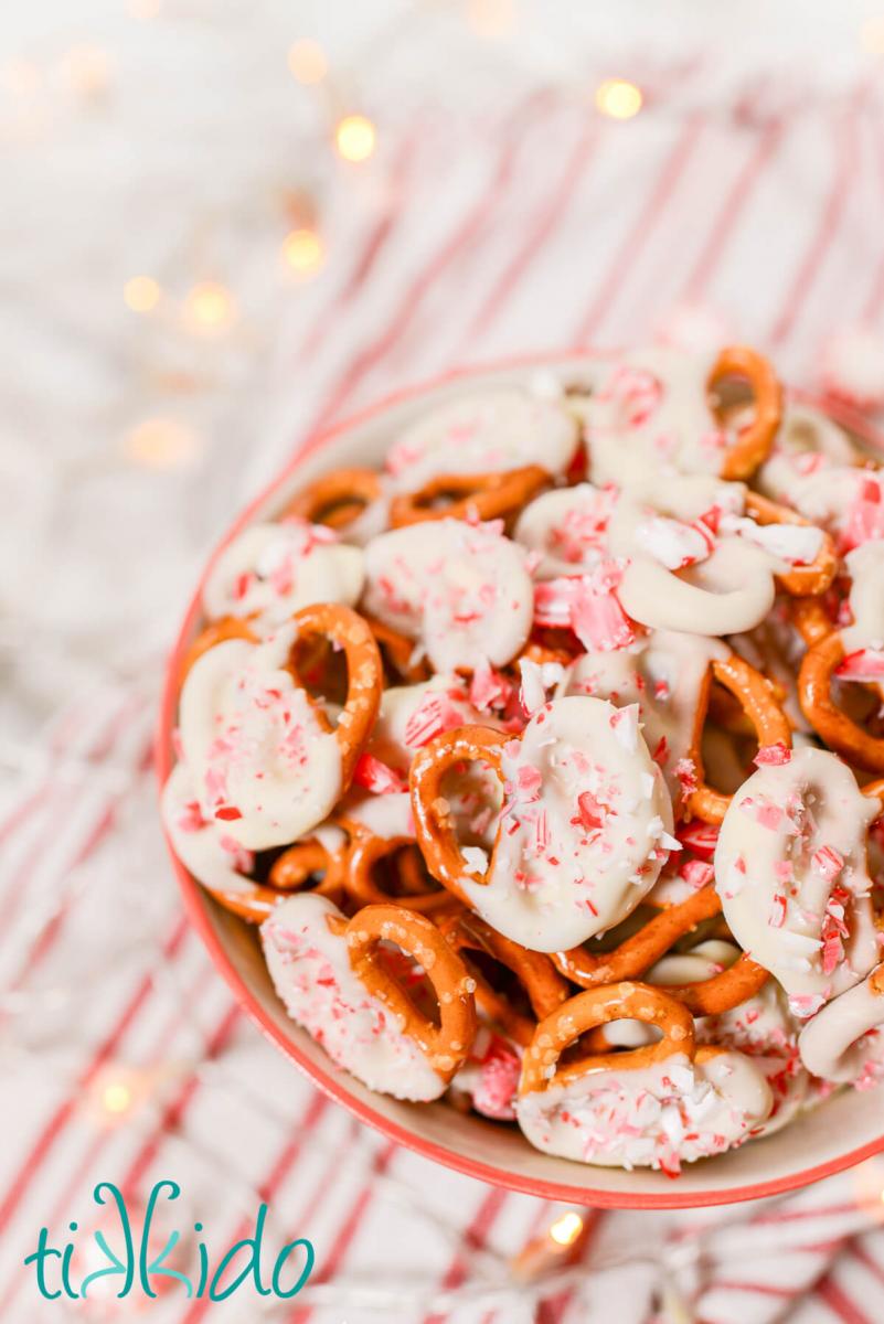 Bowl full of pretzels dipped in peppermint white chocolate and sprinkled with crushed peppermint candies.  Whole pepperming candies, twinkle lights, and a red and white striped kitchen towel surround the bowl.