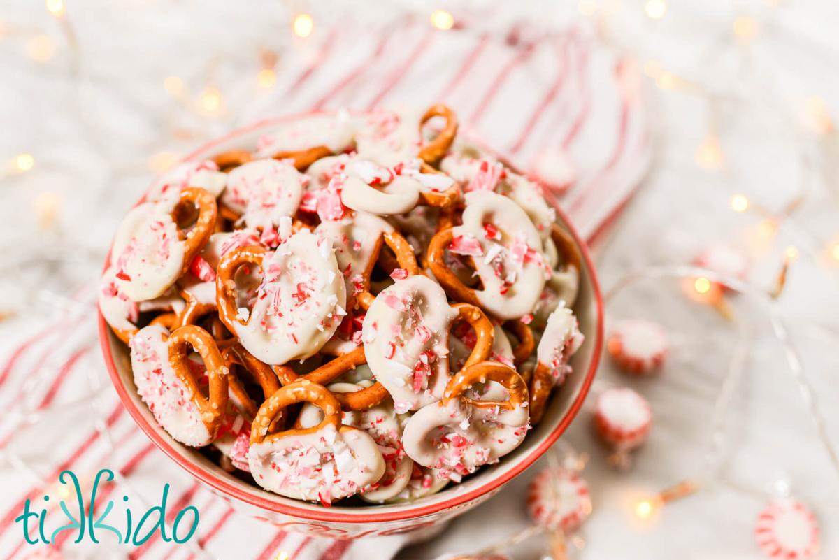 Bowl full of pretzels dipped in peppermint white chocolate and sprinkled with crushed peppermint candies.  Whole pepperming candies, twinkle lights, and a red and white striped kitchen towel surround the bowl.