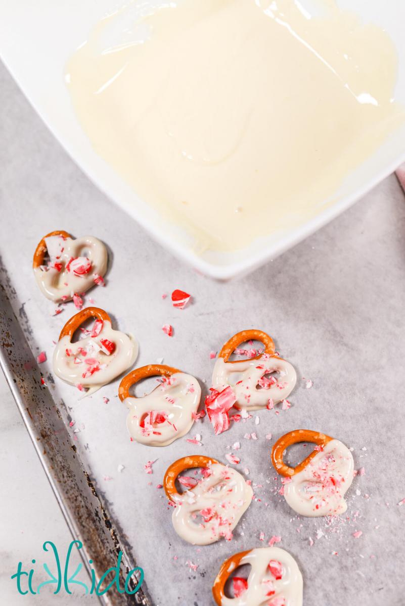 Pretzels being dipped in peppermint white chocolate, sprinkled with crushed peppermint candies, and set out to cool on a waxed paper lined baking sheet.