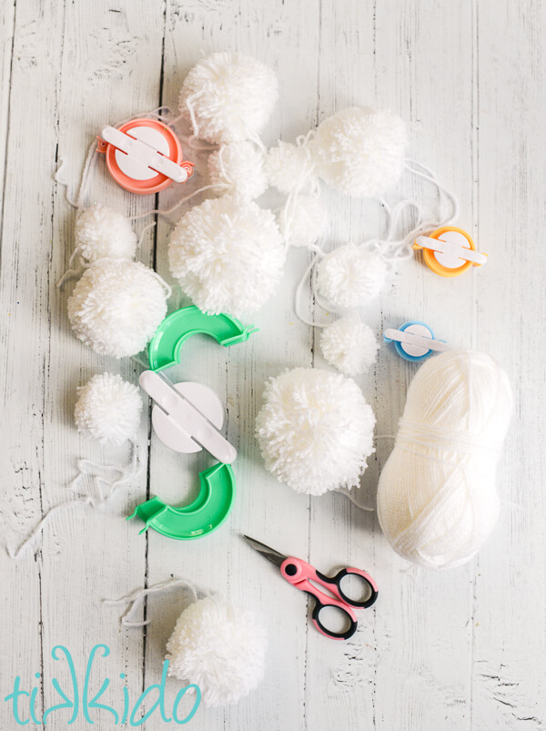 Materials for making a pom pom garland scattered on a white wooden surface.