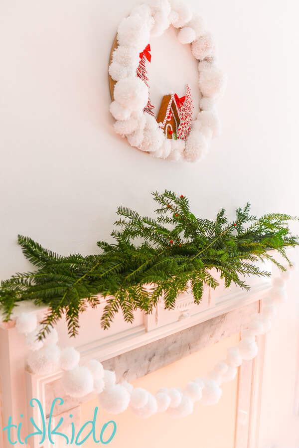 White pom pom garland hanging from a fireplace mantel decorated with fresh evergreen branches.  A pom pom christmas wreath hangs above the fireplace.
