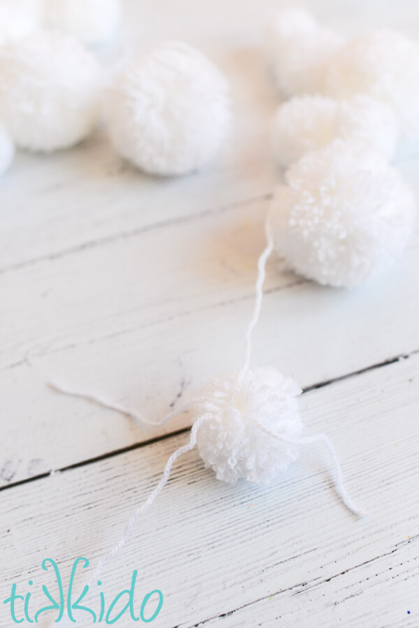 Small pom pom being tied onto a piece of yarn  showing how to make a pom pom garland.  A pile of white pom poms of various sizes are in the background.