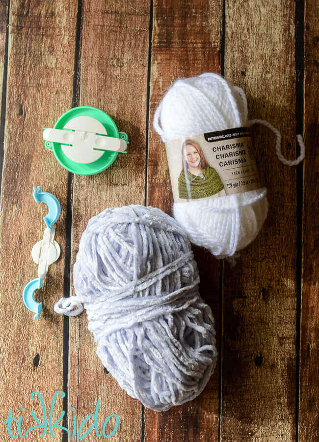 White and pale grey yarn next to two pom pom maker tools on a wooden backdrop.