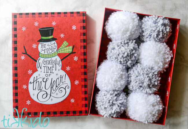 Yarn pom poms in a snowman themed gift box for a DIY Pom Pom Snowball Fight Kit