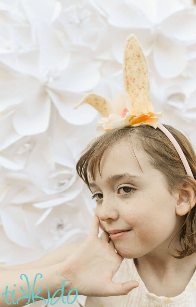 Girl wearing Bunny ear headband made with cream colored felt, a peaches and cream colored calico fabric, and ivory silk flowers on a pink headband, in front of a white surface.