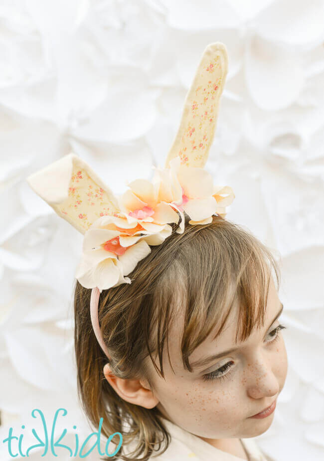 Girl wearing Bunny ear headband made with cream colored felt, a peaches and cream colored calico fabric, and ivory silk flowers on a pink headband, in front of a white surface.