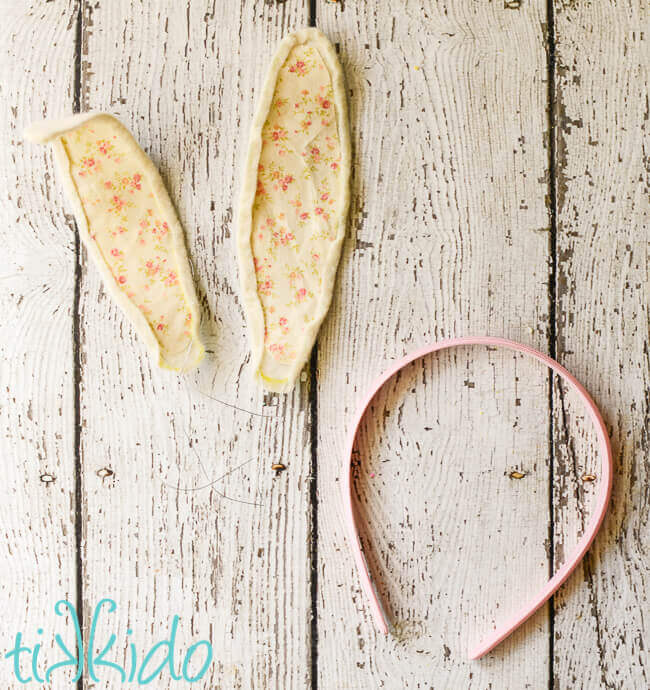 Wired fabric bunny ears next to a pink headband on a weathered, white, wooden surface.