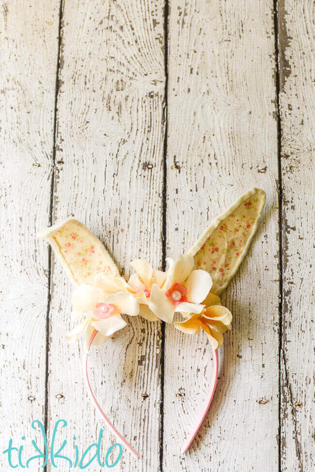 Bunny ears headband made with cream colored felt, a peaches and cream colored calico fabric, and ivory silk flowers on a pink headband, on a weathered white wooden surface.