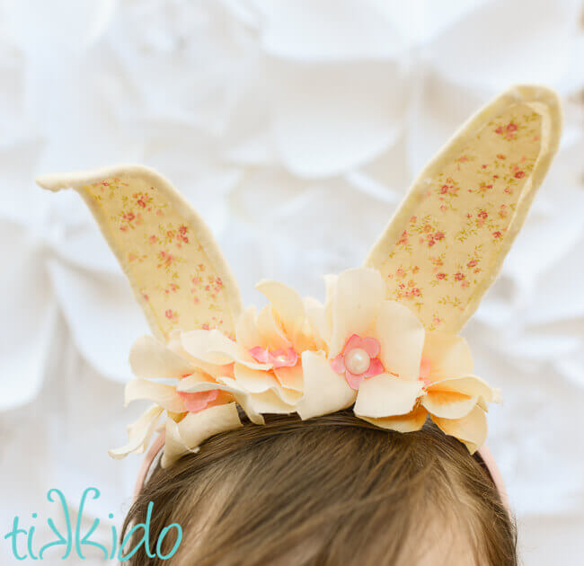 Girl wearing Bunny ear headband made with cream colored felt, a peaches and cream colored calico fabric, and ivory silk flowers on a pink headband, in front of a white surface.