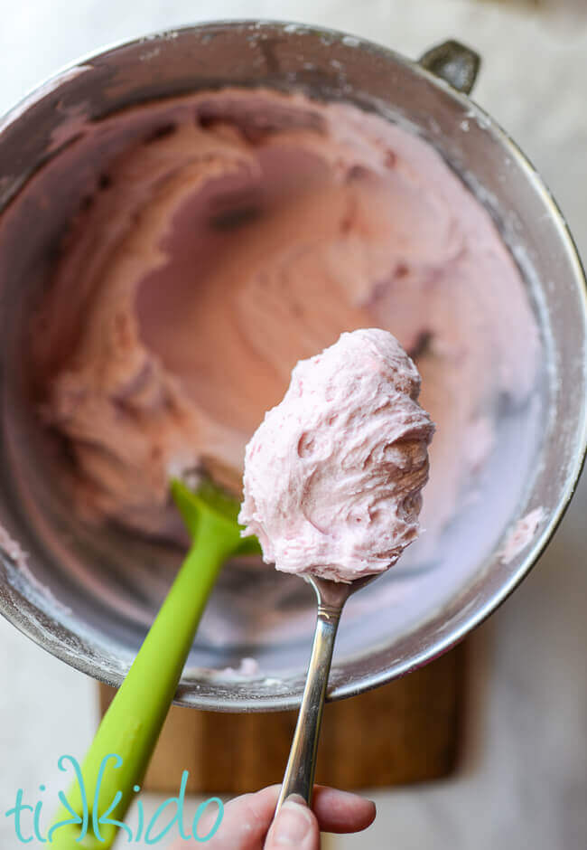 Spoon of pink prickly pear buttercream icing above a bowl of the same frosting, with a green spatula in bowl.
