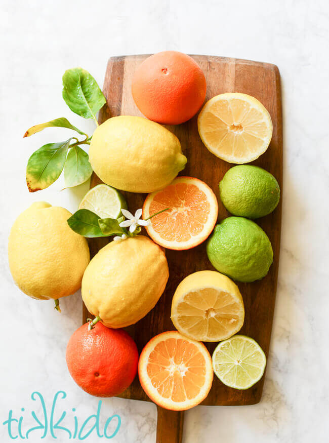 Fresh oranges, lemons, and limes cut and whole on a wooden cutting board on a white marble surface.