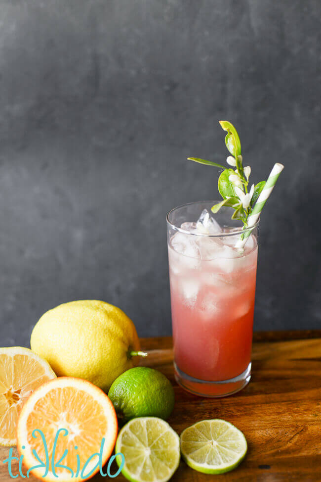 Pink Prickly pear margarita in a tall glass with ice and a green striped paper straw embellished with fresh citrus blossoms.