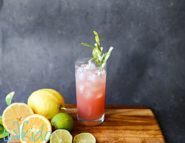 Pink Prickly pear margarita in a tall glass with ice and a green striped paper straw embellished with fresh citrus blossoms.