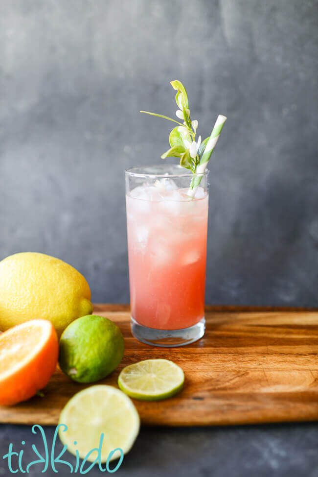 Pink Prickly pear margarita in a tall glass with ice and a green striped paper straw embellished with fresh citrus blossoms.