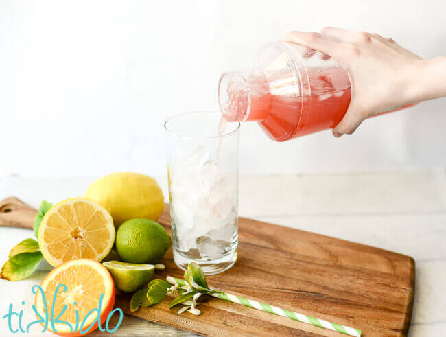 Pink prickly pear margarita being poured from a clear cocktail shaker into a tall glass.  Lemons, limes, and oranges sit beside the glass.