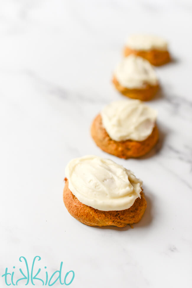 Soft pumpkin cookies with cream cheese frosting in a line on a white marble table.