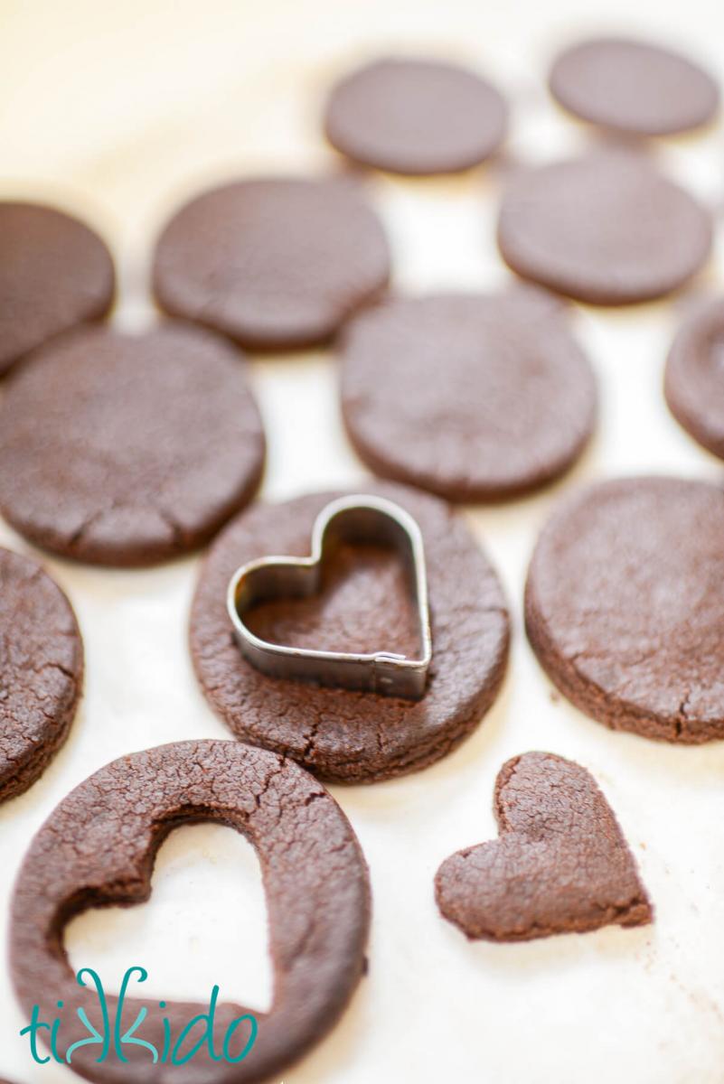 Chocolate cut out cookies baked in round shapes, with a heart shaped cookie cutter being used to cut a heart shaped window in some of the chocolate cookies.