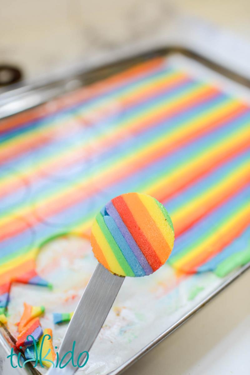 A circle of rainbow frosting being lifted away from a sheet pan with an offset spatula, ready to be assembled into a homemade rainbow oreo.