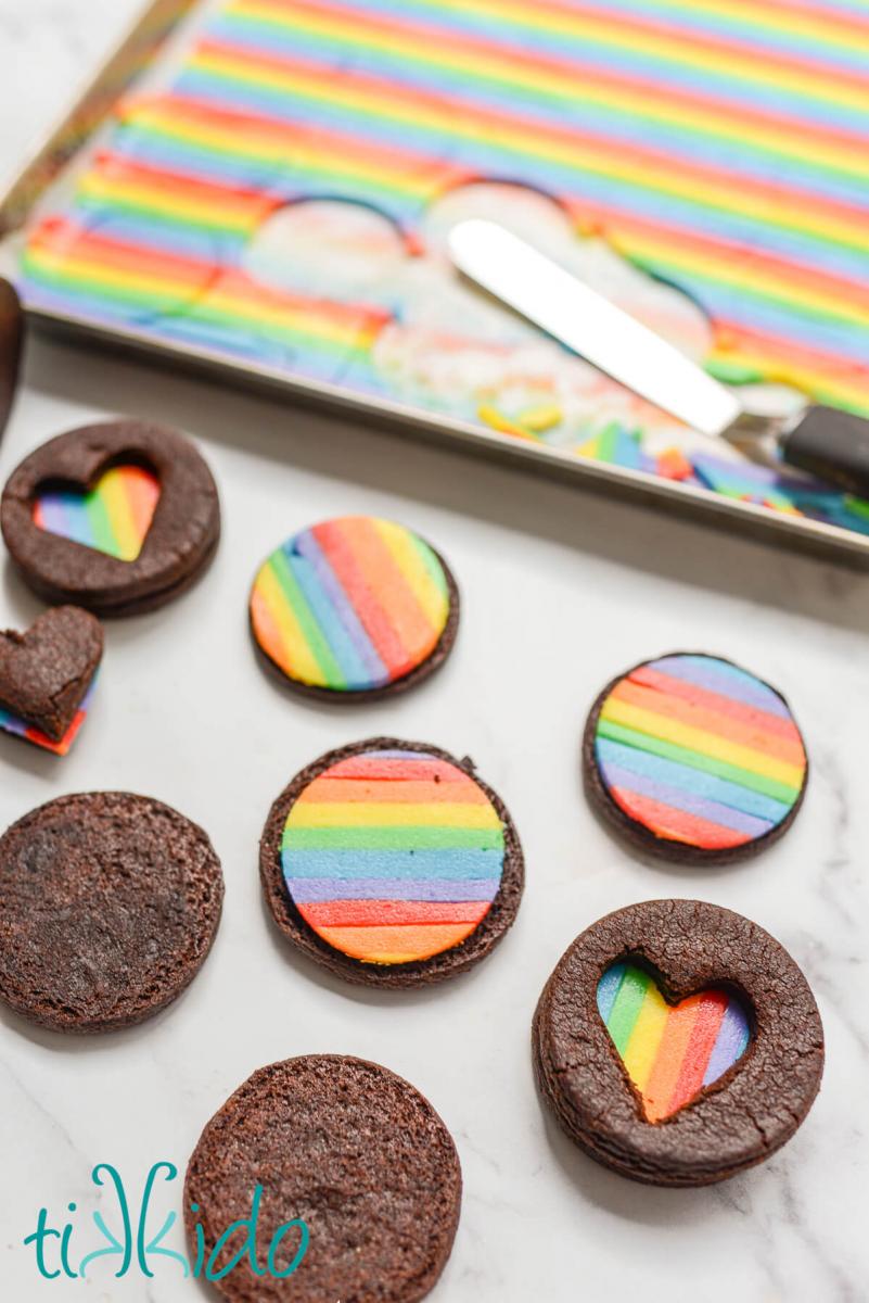 Homemade rainbow oreos be filled with rainbow frosting centers.  A sheet pan with icing piped in rainbow stripes sits behind the rainbow sandwich cookies being assembled.