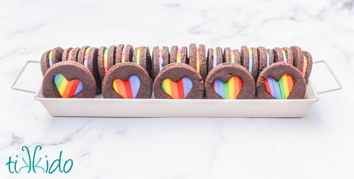 Homemade rainbow oreos with heart shaped cut outs arranged in a white rectangular tray.