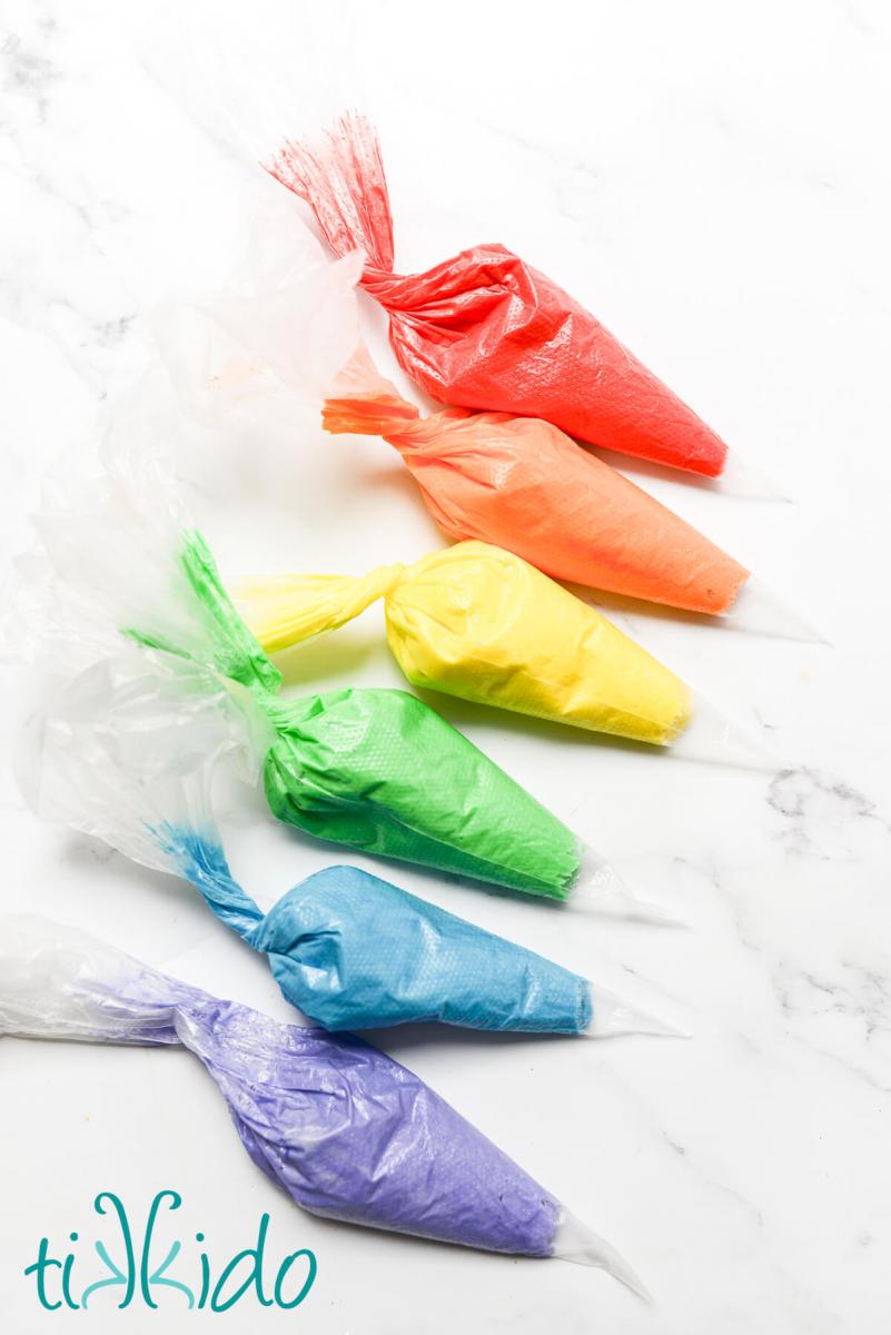 American buttercream icing colored in rainbow colors, in disposable pastry bags.  The pastry bags sit on a white marble surface, lined up in the colors of the rainbow.