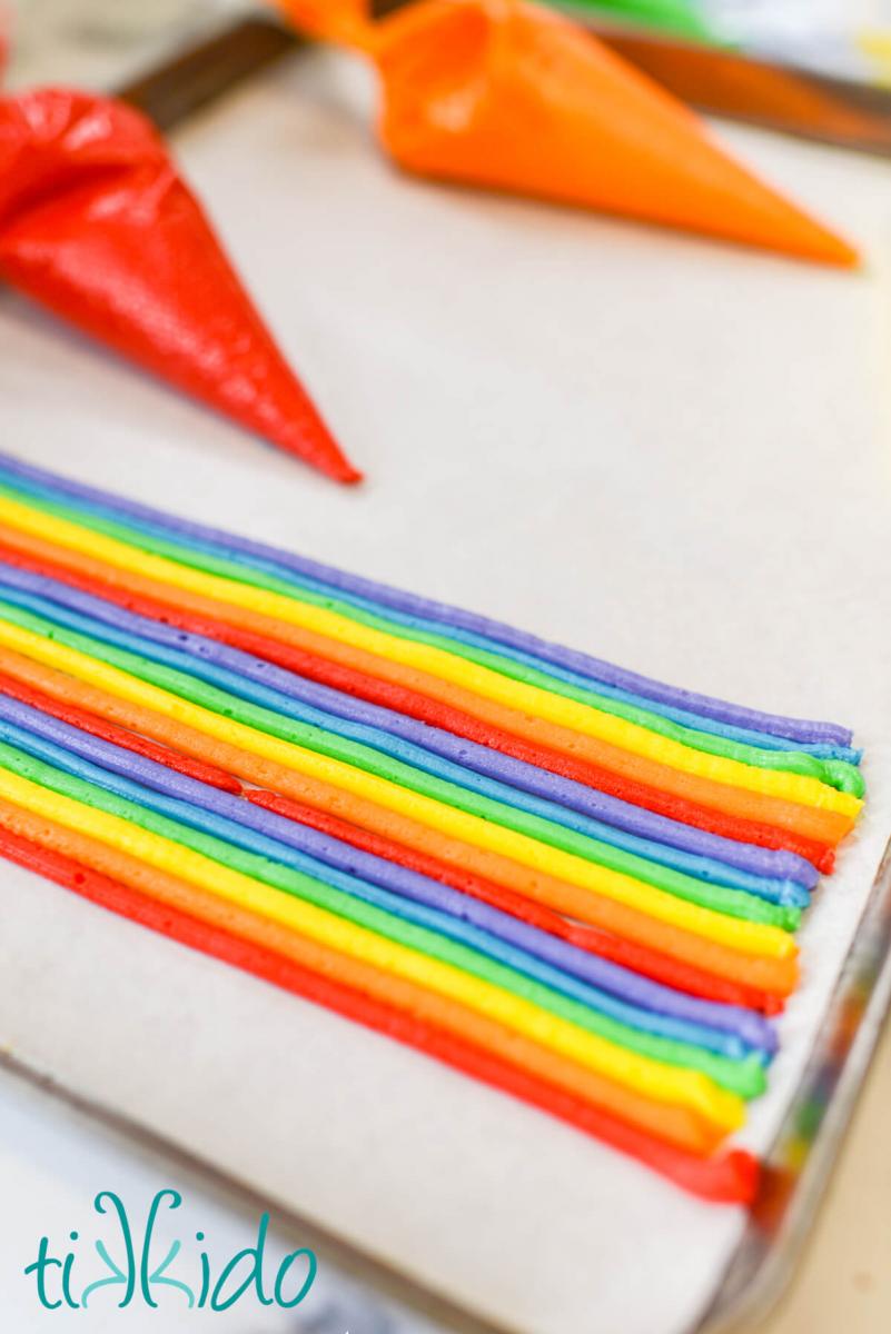 American buttercream frosting being piped into rows in the order of the rainbow.  The frosting is piped on parchment paper sitting on a sheet pan.