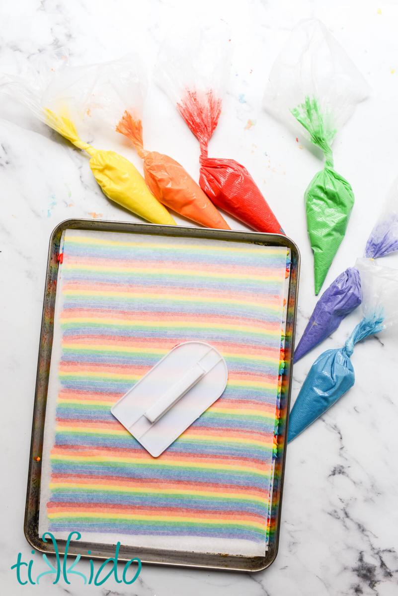 Piped rows of rainbow frosting with a sheet of parchment paper on top, being smoothed with a fondant smoother.