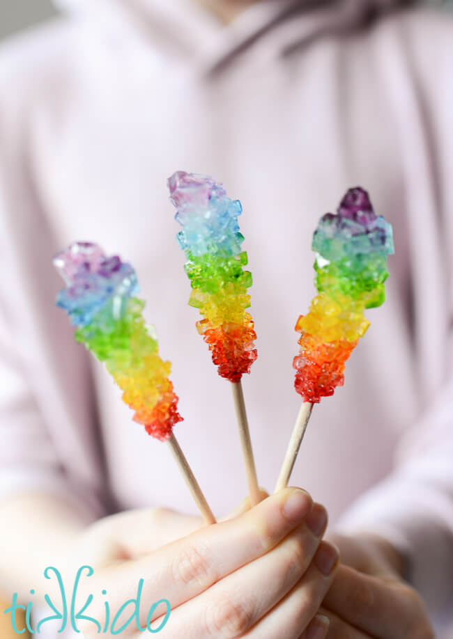 Girl holding three sticks of rainbow rock candy in her hand.