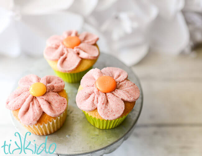 Three vanilla cupcakes on a galvanized metal cake stand.  The cupcakes are iced to look like pink flowers with five petals.