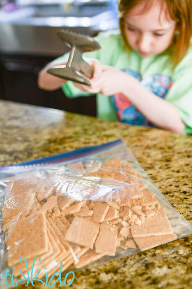 Little girl using a meat tenderizing mallet to crush graham crackers in a gallon sized ziplock bag.