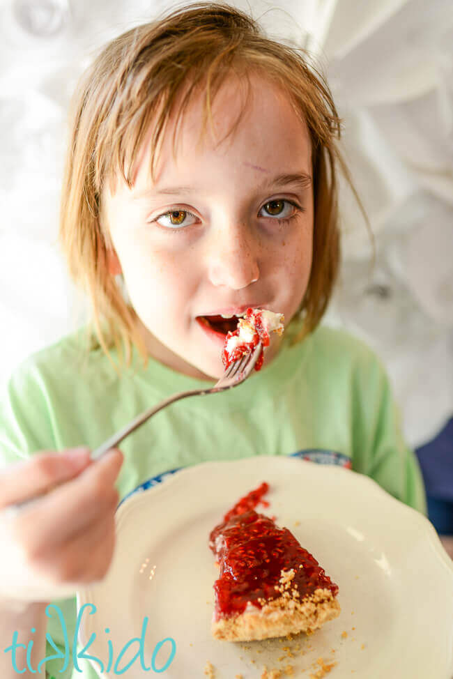 Little girl with messy hair eating a bite of raspberry cream pie against a white floral backdrop.