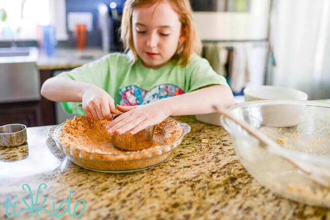 Little girl using a metal cup measure to press graham cracker crumbs into a pyrex pie plate.