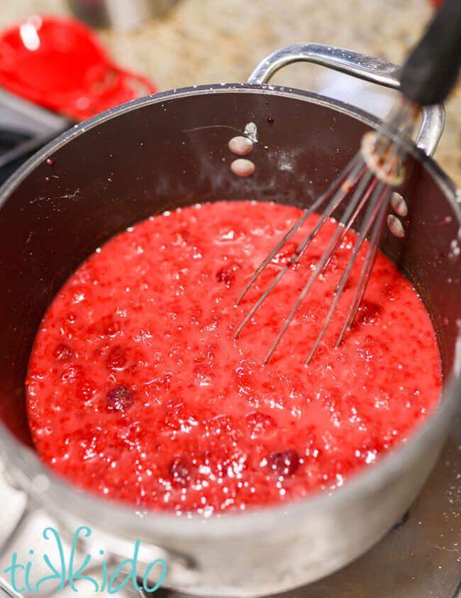 Raspberry layer of a raspberry cream pie being cooked in a 4 quart saucepan on a gas stove, being stirred with a whisk.