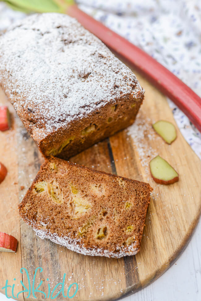 A sliced loaf of rhubarb bread on a wooden cutting board.  Slices of fresh rhubarb and a stalk of rhubarb are scattered beside the quick bread.