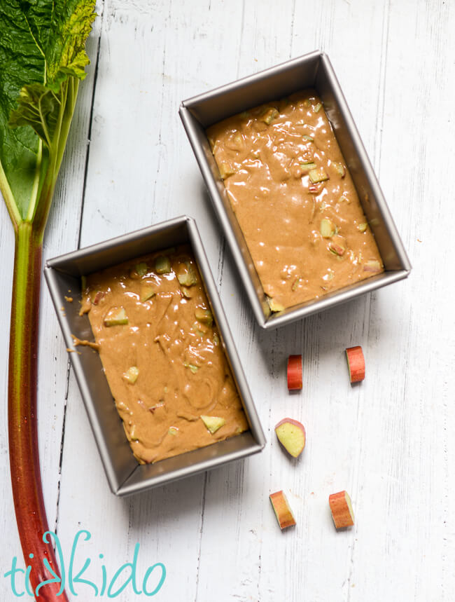 Batter for rhubarb bread divided between two metal bread loaf pans sitting on a white wooden surface.  A fresh stalk of rhubarb and rhubarb slices surround the pans.