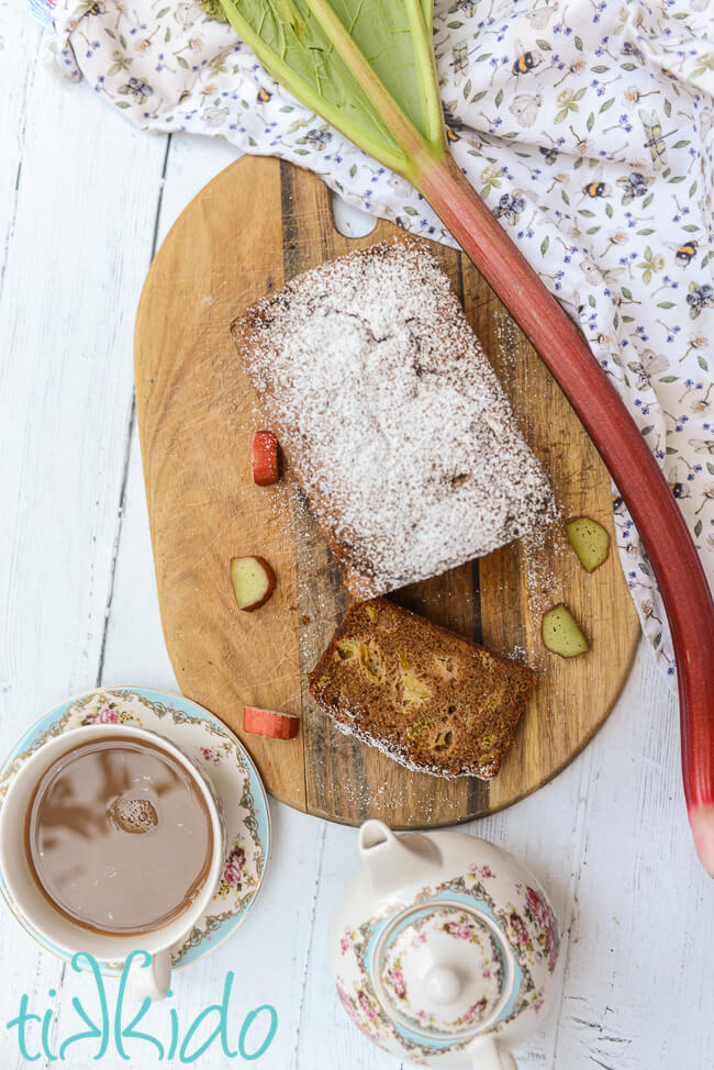 A loaf of rhubarb bread with one slice cut, on a wooden cutting board, surrounded by a cup of tea and small teapot, slices and a stalk of rhubarb, and a springy tea towel.