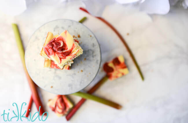 Rhubarb cheesecake bars on a metal cake stand, surrounded by stalks of fresh rhubarb.