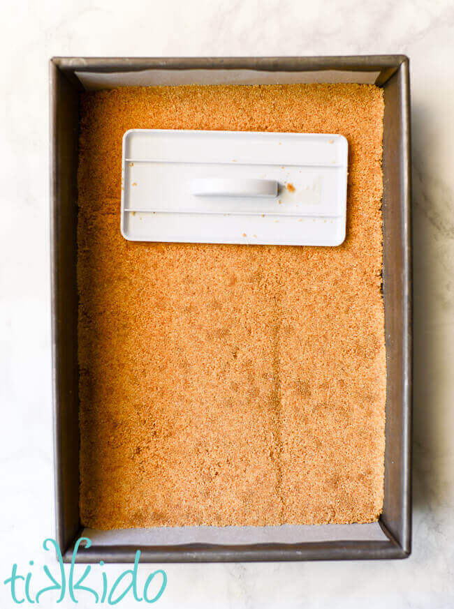 Graham cracker crust being pressed into a 9x13 pan using a fondant smoother.