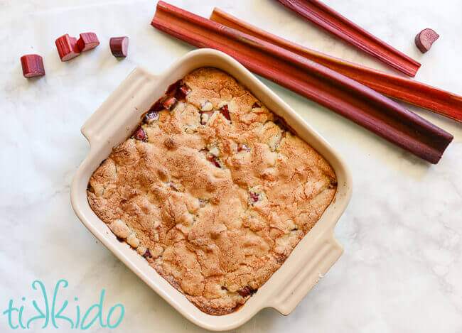 rhubarb cobbler in an 8x8 baking dish on a white marble surface, surrounded by slices and stalks of fresh rhubarb.