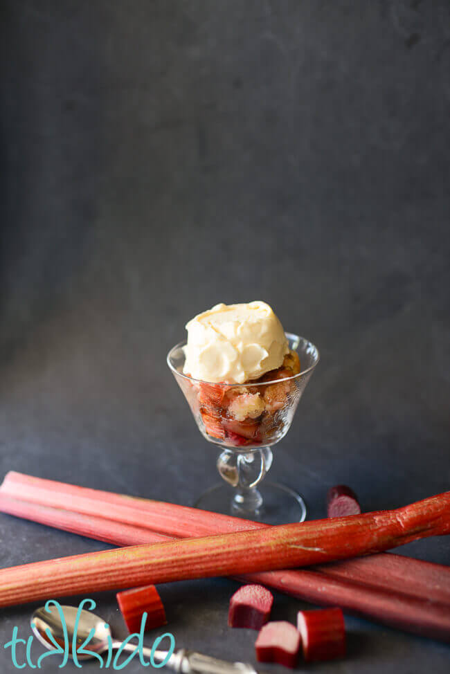 Rhubarb cobbler in a glass serving dish, topped with vanilla ice cream.  The glass sits on a black background, and is surrounded by stalks and slices of fresh rhubarb.